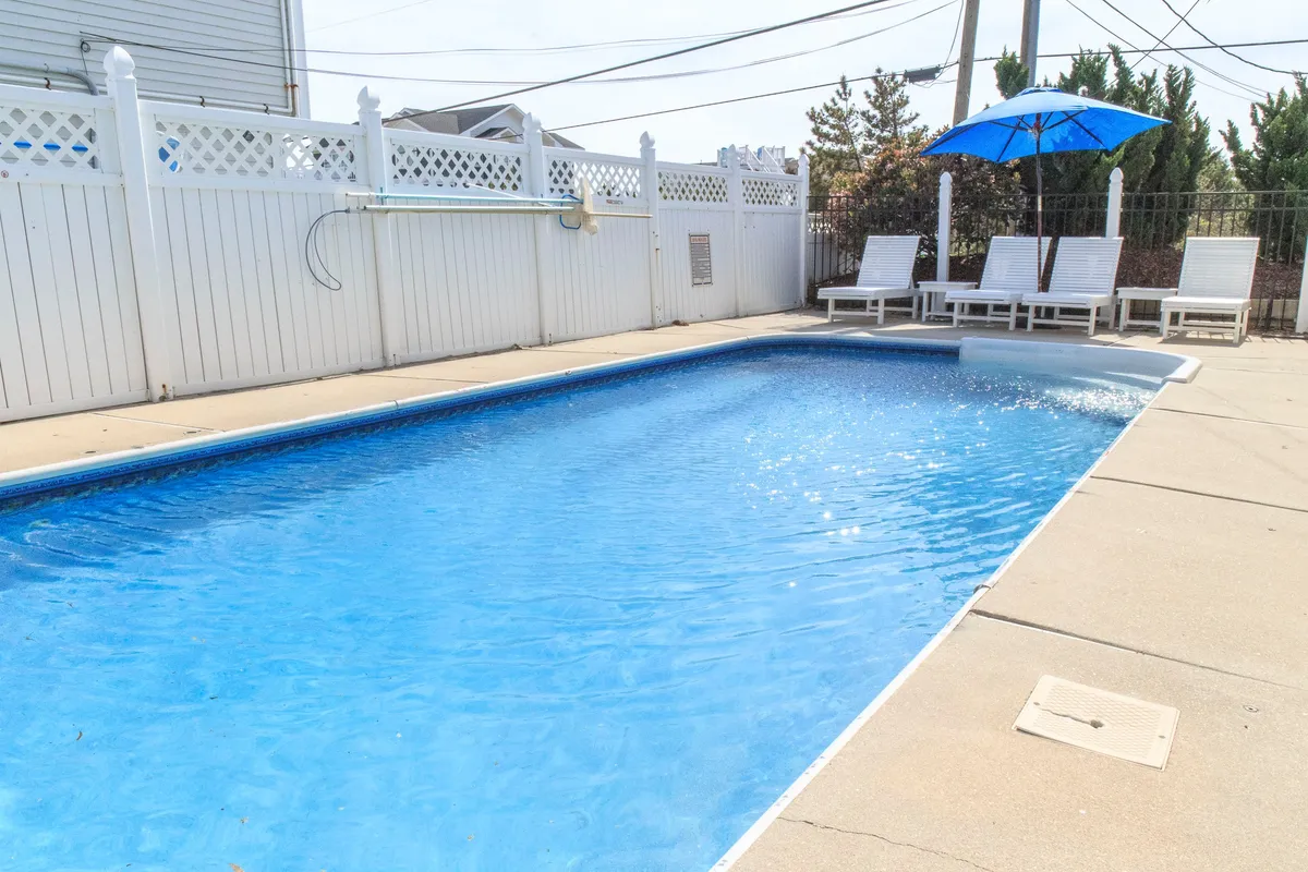 The image showcases the exterior of a Mesa, Arizona single-family house, centered around a sparkling swimming pool flanked by inviting lounge chairs and a vibrant blue umbrella. The scene exudes a leisurely atmosphere perfect for relaxing, sunbathing, swimming, or gathering for sunset swims and poolside barbecues.