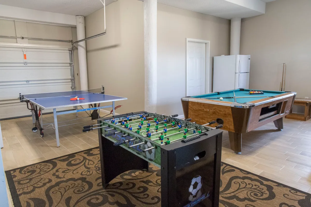 A spacious interior game room featuring a ping pong table, foosball table, pool table, and refrigerator. The room is designed for fun gatherings and includes a visible bar area that could double as bar seating, with the refrigerator hinting at kitchen adjacency.