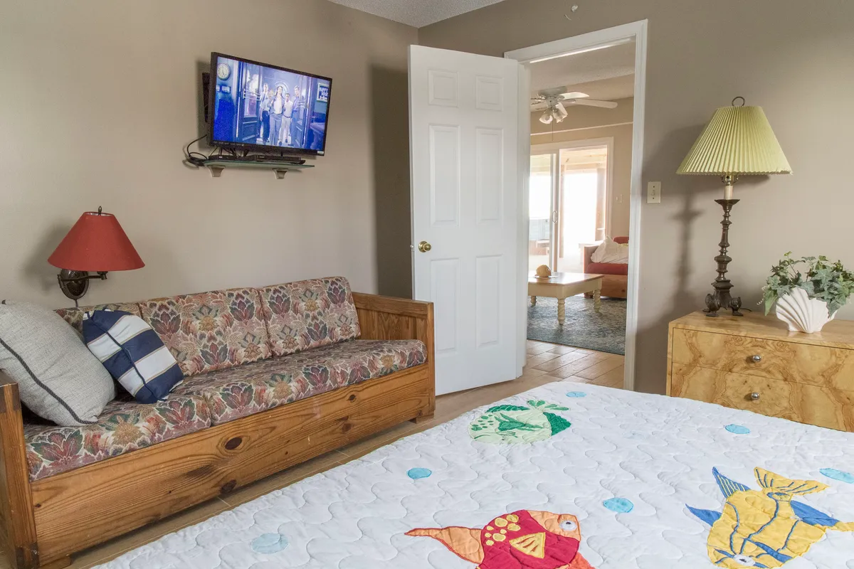 A cozy living area featuring a quilted bedspread with playful fish designs, a sofa, television, a lamp, and a doorway leading to another room. The scene exudes warmth and comfort, ideal for relaxed movie nights, reading, or quiet family time.