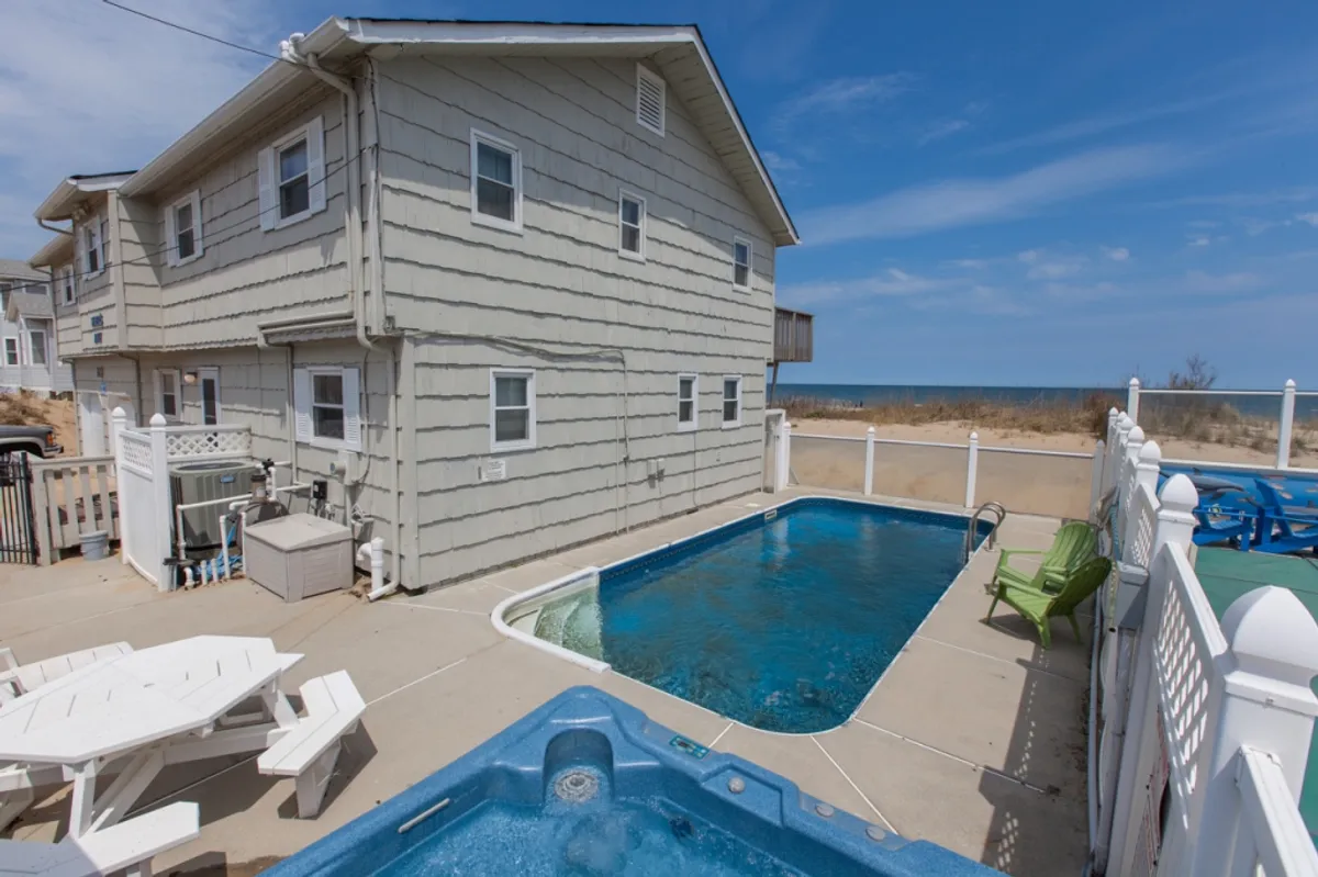 The scene features a spacious outdoor area with a swimming pool, hot tub, and dedicated seating area. The backyard is designed for relaxation and entertaining, offering ample space for barbecues or quiet enjoyment. While the setting includes a beach view in the distance, the focus remains on the usability and comfort of the backyard itself.