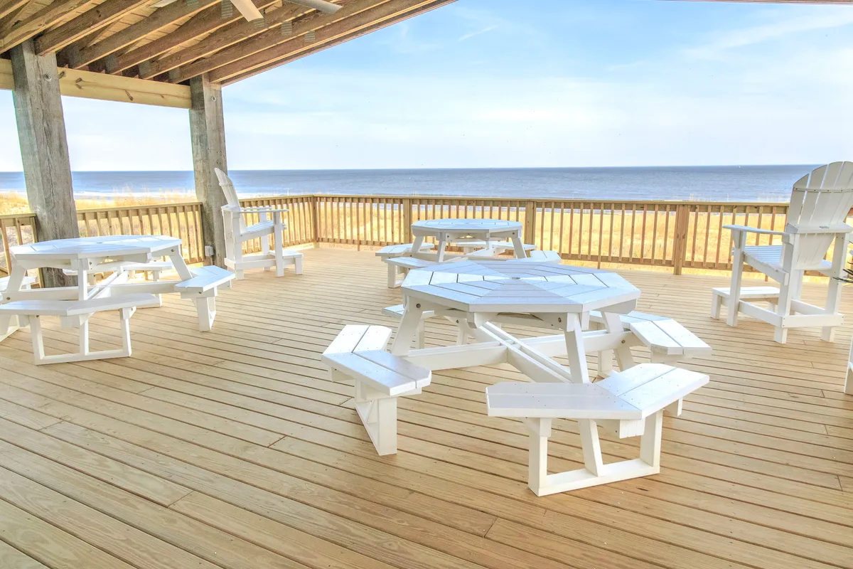 A spacious wooden deck furnished with a distinctive octagonal picnic table and white Adirondack chairs, overlooking a serene ocean backdrop. The scene suggests ample room for families to gather, dine, play games, or simply enjoy the fresh air and beautiful view. The atmosphere is casual and joyful, inviting togetherness and outdoor fun.
